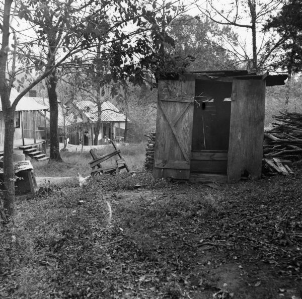 A building, possibly an outhouse, with a woodpile behind it. In the background, two houses with porches are visible. "No roads or streetlights or sidewalks or sewers. Only wood piles and ragged outhouses."<p>80 people live in 21 buildings in the Taylor Alley neighborhood of Natchez. The occupants have 11 hydrants, two indoors, and 15 toilets.