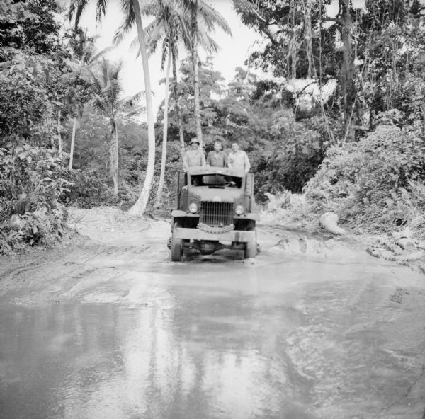 Three soldiers in a truck drive through mud on Kiriwina Island in the Solomon Sea, New Guinea (present day Papua New Guinea). Jungle foliage is in the background.