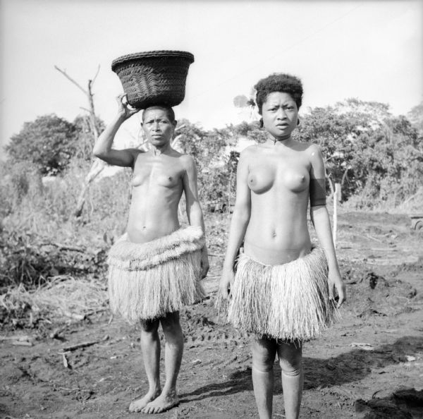 A young indigenous woman wearing a flower in her hair poses with an older woman who is balancing a basket on her head on Kiriwina Island in the Solomon Sea, New Guinea (present day Papua New Guinea). Their only garments are grass skirts. The jungle is in the background. Robert Doyle wrote a caption for this image although it was not published at that time, "Yams are one of the principal items in the diet of Kiriwina natives. Yams are somewhat similar in shape and taste to sweet potatoes. They are carried home from the famous native gardens of Kiriwina in baskets of the type shown in the photograph. American soldiers and war correspondents often stop native women to take pictures of their yam baskets."