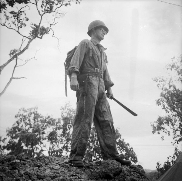 Bob Eunson of Associated Press at Port Moresby, New Guinea (present day Papua New Guinea), dressed in a "combat suit." He is wearing a pack on his back and carries a machete.