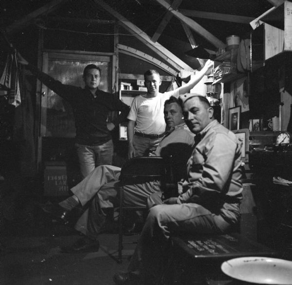 The interior of the "Sad Shack" at Camp Cable, near Brisbane, Australia. Two men are standing and two men are sitting in chairs. Personal items such as portraits and pin-ups decorate the walls. Handwritten on the reverse of the print, "Sad Shack gang, Elmer Boyle, Redd Allen, Bob Gillette, ?, 1943, Camp Cable, Brisbane."