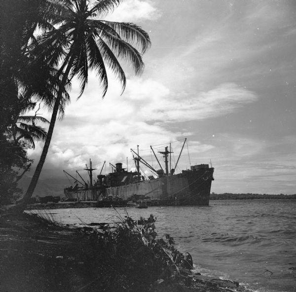 A warship moored at the Island Jetty Dock on Goodenough Island, in the Solomon Sea, New Guinea (present day Papua New Guinea). A palm tree is silhouetted on the left. Trucks are on the dock, mountains are in the background.