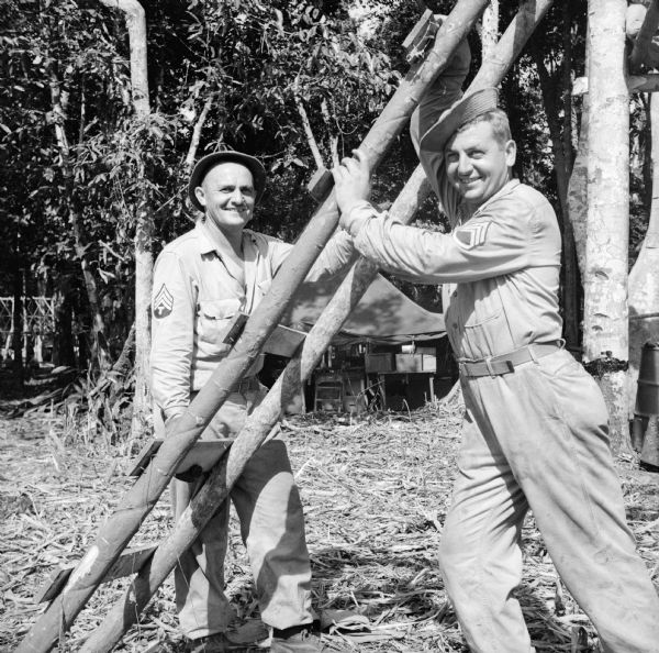 A rickety ladder serves (Sergeant) Technician 4th Grade John Duray (left), and his brother, Staff Sergeant Isador Duray, both of Elkhorn, Wisconsin, in the construction of a camp on Goodenough Island in the Solomon Sea, New Guinea (present day Papua New Guinea). They were in the same unit for two and a half years. Tents and trees are in the background.