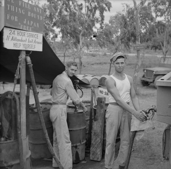 Corporal Gareale Brezeale of Warren, Arkansas, stands next to barrels, and Private First Class William Imhoff of Detroit, Michigan, holds the nozzle to the gas pump at the Kangaroo Korner Gas Station near the entrance to the 5th Air Force Headquarters near Port Moresby, New Guinea (present day Papua New Guinea). Signs overhead read, "We Fill Anything Bound For Japan For 'Free'" and "24 Hour Service, If Attendant Isn't Around, Help Yourself." A jeep and trees are in the background.