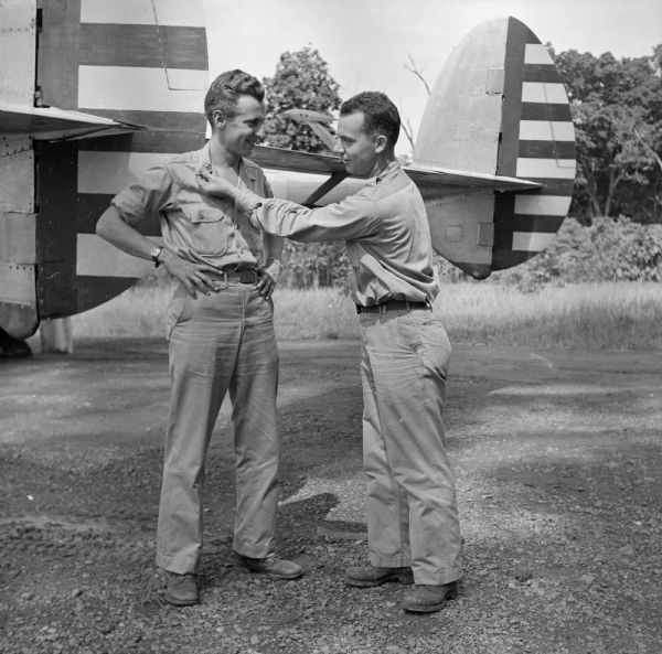 Robert Doyle admires the golden oak leaf of Major Post's collar insignia. They are standing in front of the tail assembly of a plane similar to Post's. Major Arthur L. Post of Milwaukee, Wisconsin, was shot down on June 20th, 1943 and rescued on September 28th, 1943, after 101 days in the jungle. The airfield where this image was taken was located near Port Moresby, New Guinea (present day Papua New Guinea). Robert Doyle was not allowed to publish Post's story at that time. It was finally published in January, 1947, by the <i>Milwaukee Journal</i>. Major Post intended to write his own story, but was killed in a test airplane on August 25th, 1944, before he had the chance.