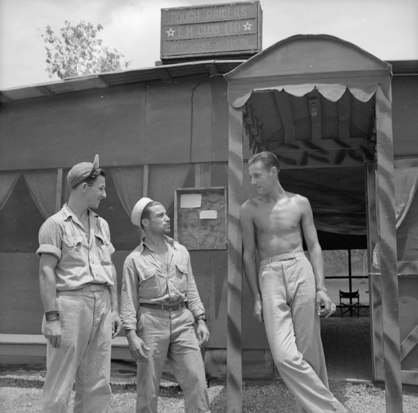 Motor Pool Dispatchers (left to right) Sergeant Alex Petro of Racine, Wisconsin, and Sergeant Joe Nicolazzi of Kenosha, Wisconsin, chat with Corporal Eugene Tobbs (shirtless) of Pontiac, Michigan, a custodian taking a break in front of the Rough Raiders Enlisted Men's Club, Ltd. The sign advertises "Refreshments - Recreation."