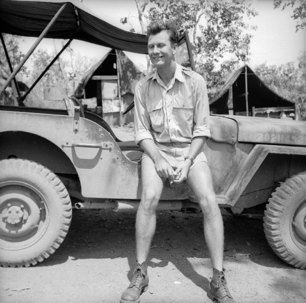 Navigator Lieutenant Fred Radtke of Milwaukee, Wisconsin, poses while perched on the side of a jeep. The base is near Port Moresby, New Guinea (present day Papua New Guinea). Tents are in the background.