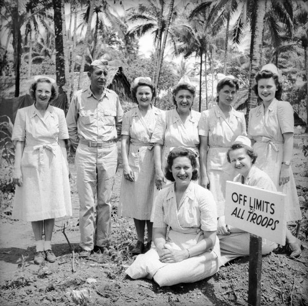 Seven Lieutenant nurses from Wisconsin pose with a Lieutenant Colonel from Illinois behind a sign that declares "Off Limits To All Troops." Names (left to right, standing) are Lieutenant Marie Kuenzi of Beaver Dam, Lieutenant Colonel Max C. Ehrlich of Chicago, Lieutenant Marianna Brown of Beloit, Lieutenant Phyllis Koll of Milwaukee, Lieutenant Doris Gregory of Shorewood, and Lieutenant Geraldine Buergin of Madison. Seated (left to right) are Lieutenant Doris Grube of Kenosha and Lieutenant Muriel Cannon of Ellsworth. Buildings and palm trees are in the background. The location is on Milne Bay, New Guinea (present day Papua New Guinea).