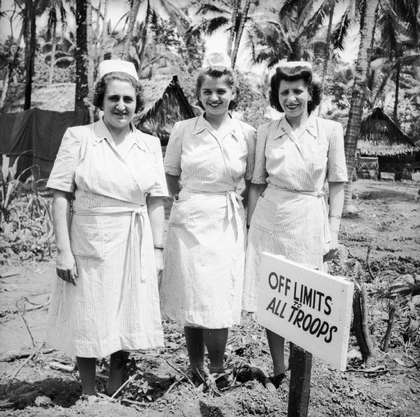 Three Lieutenant nurses from Michigan pose behind a sign that declares "Off Limits To All Troops." Names, left to right, are Lieutenant Avice Cleveringa of Grand Haven, Lieutenant Irene Koehn of Detroit, and Lieutenant Ruth Holy of Battle Creek. Buildings and palm trees are in the background.