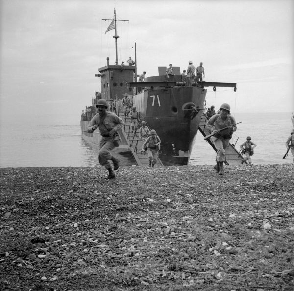 32nd Division Infantry Combat Team landing from LCI (Landing Craft Infantry) in a practice exercise. Soldiers are running down a ramp, then up a rocky beach from the landing craft. The number on the ship is "71." The ship is flying the American flag. Robert Doyle wrote a caption for this image although it was not published at that time: "Infantrymen who landed at Saidor, New Guinea, run off LCI in exercise few weeks before actual landing."
