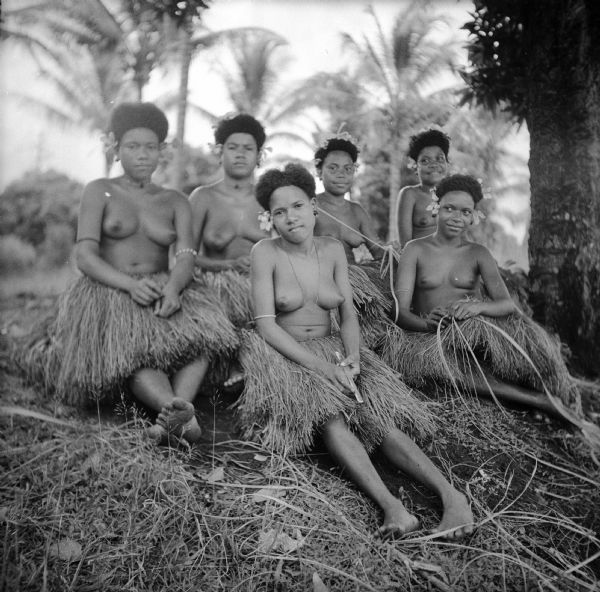 A group of six young indigenous women posed seated on the ground. They are wearing grass skirts and flowers in their hair. The woman in front is holding a knife. The location is near Milne Bay, New Guinea (present day Papua New Guinea).