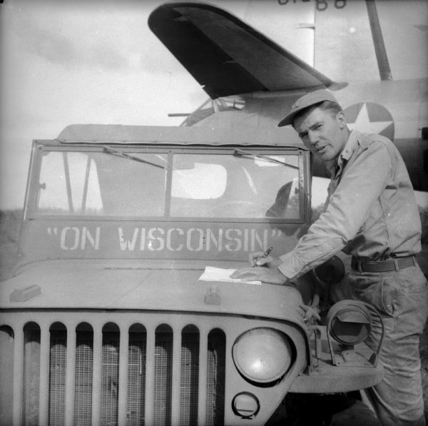 Ordnance Lieutenant Elroy Derksen of Milwaukee, Wisconsin, glances up from writing on papers laying on the hood of his jeep at a military base in New Guinea (present day Papua New Guinea). His jeep has the phrase "On Wisconsin" painted below the windshield. "On Wisconsin" is the title of the University of Wisconsin Fight Song and the State Song of Wisconsin. The tail of a warplane is behind him.