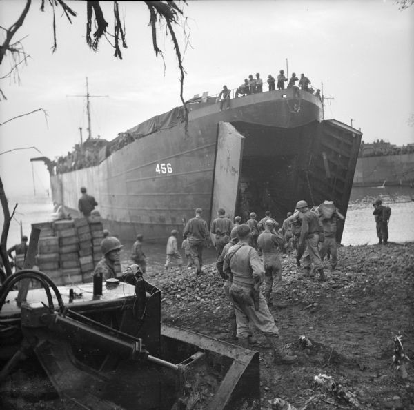 LST (Landing Ship, Tank) on the beach at Saidor, New Guinea (present day Papua New Guinea). Soldiers are unloading vehicles and supplies for the Saidor landing. A bulldozer is on the left and another LST is on the right. Robert Doyle's caption reads: "Few minutes after ships hit rocky beach at Saidor, trucks and jeeps race along beach and into jungle. Big elevator on ship lowers vehicles from top deck to hold. they are then driven out open bow of ship."