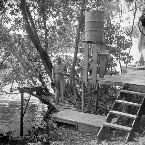 A soldier is standing on the ramp leading to the camp toilet suspended over the water, with a roll of toilet paper sitting on the edge. On the right is a water barrel on a platform with a shower head. Up four wooden steps to a platform is a wash basin on the other side of the water barrel. Towels are on the far right. Trees on the shore offer some privacy. The location is Dreger Harbor on the north coast of New Guinea (present day Papua New Guinea). Note on the back of the print, "Shower and one holer."