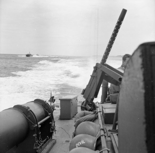 View of two PT (Patrol Torpedo) boats from another PT boat, off of the north coast of New Guinea (present day Papua New Guinea). PT (Patrol Torpedo) boats were a variety of torpedo-armed fast attack craft used to attack larger surface ships. One of the boat's guns and some of the soldier's gear and helmets can be seen on the deck. A soldier is sitting (partially obscured) on the right.