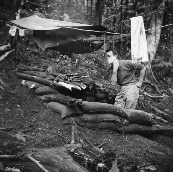 Robert Doyle shaves while looking in a mirror suspended from a hammock with a rain tarp in the military camp at Saidor, New Guinea (present day Papua New Guinea). Sand bags are stacked to create an alcove, and his helmet is hanging from a branch, probably full of water. A towel is hanging on the right, and clothes are stacked on the left.