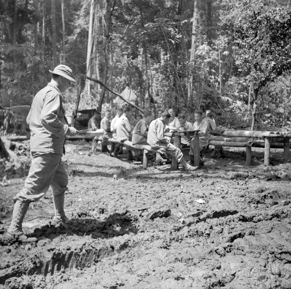 Officers sit down together for a meal at the Task Force Headquarters Mess in the military camp at Saidor, New Guinea (present day Papua New Guinea). One soldier is walking through the rutted mud in the foreground. The jungle is in the background. Robert Doyle wrote a caption for this image although it was not published: "The Elite of the Saidor task force dine here. This is the headquarters officers' mess. They stand in the chow line with mess kits and carry their food to the rough pole tables. When it rains at mealtime the elite get rained on."