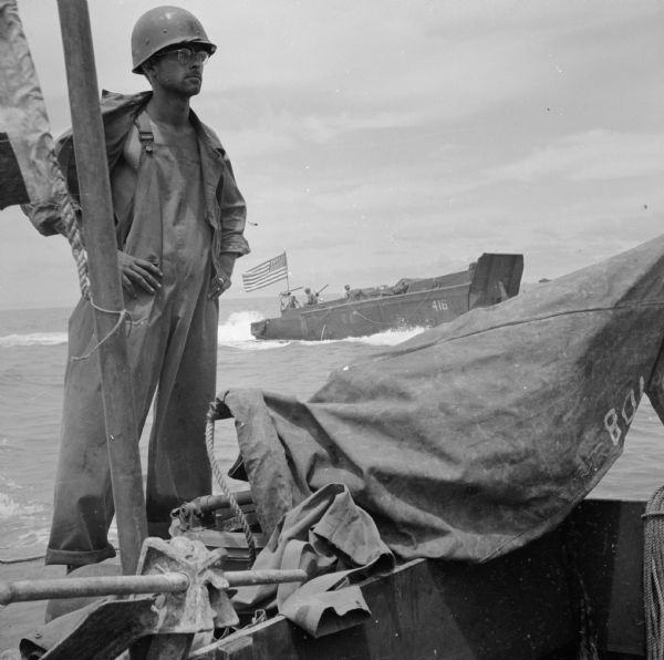 Corporal Andrew Starkie of Chicago, Illinois, stands next to the flagpole on a LCV (Landing Craft, Vehicle) off of the coast of Saidor, New Guinea (present day Papua New Guinea). Another LCV, flying the American flag, is in the background.