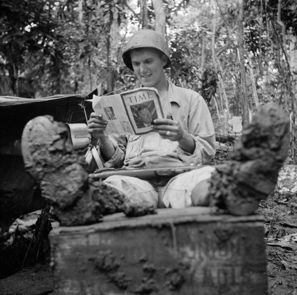 A soldier, Sergeant Robert Witzlsteiner of Milwaukee, Wisconsin, reads <i>TIME</i> magazine with his very muddy boots propped on a box. His rifle is across his lap. The military camp is at Saidor, New Guinea (present day Papua New Guinea). The jungle is behind him, and his hammock and gear appear on the left.