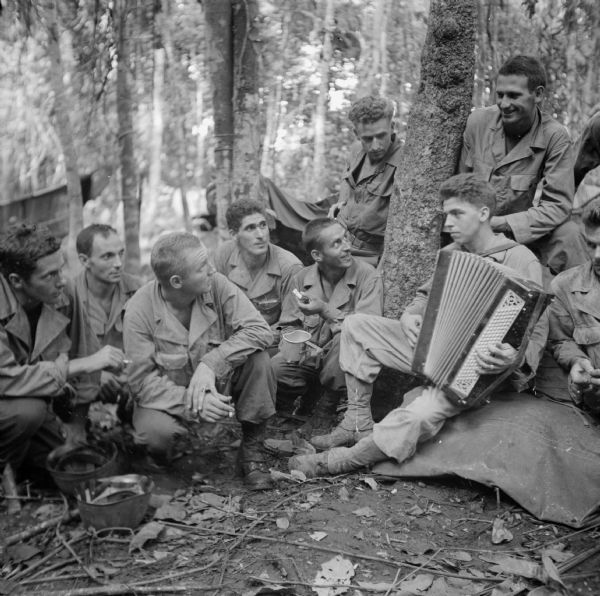 Sergeant Ed Gigowski of Milwaukee, Wisconsin, plays his accordion for some members of the 121st Field Artillery Battalion at Saidor, New Guinea (present day Papua New Guinea). They are surrounded by trees.