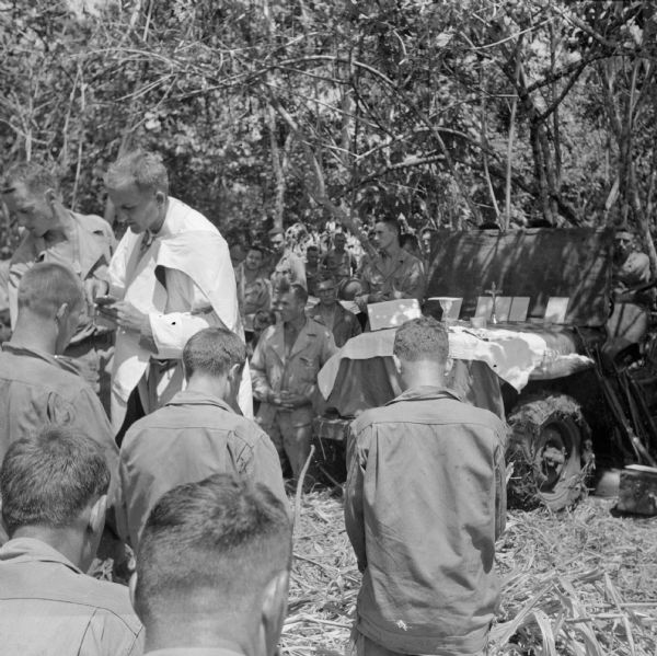 Catholic Mass at Saidor, New Guinea (present day Papua New Guinea). The Chaplain is using the hood of a jeep as an altar. Many soldiers surround the jeep as they worship. Trees are in the background. Robert Doyle wrote a caption for this image although it was not published at that time: "Army Jeep serves as altar at Catholic mass conducted by Father Anthony E. Burakowski, 30, Fairfield, Conn., Jan. 23 at artillery headquarters area at Saidor, New Guinea. Lieutenant Burakowski arrived at area in 'grasshopper' observation plane. It was his first plane ride. He arrived in New Guinea from America in December, 1943."