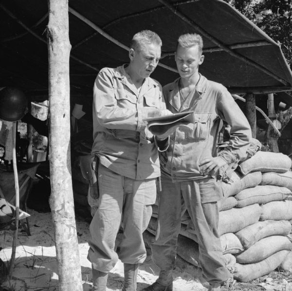 Two Wisconsin servicemen, Lieutenant Colonel Harvey W. Storm of Merrill and Private First Class Dorrison Buros of Viroqua, look at a notebook at the military camp at Saidor, New Guinea (present day Papua New Guinea). They are standing in an area with a tarp covered roof and sandbags.
