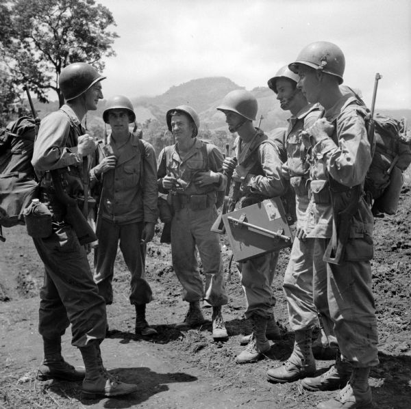 Artillerymen prepare to leave their camp in Saidor, New Guinea (present day Papua New Guinea) on a patrol that would last several days. They were to search for enemy targets against which they would direct fire by their portable radio. Names (left to right) are Captain Earl Gantenbein of LaCrosse, Private Vernon Toutant of Chippewa Falls, Private Clifford Olin of Eau Claire and Sergeant Leslie Michaud, all from Wisconsin, Private Forrest Sappington of Southwest City, Missouri and Corporal Bill Reiter of Chippewa Falls, Wisconsin. They are wearing combat gear and carrying weapons and a radio.