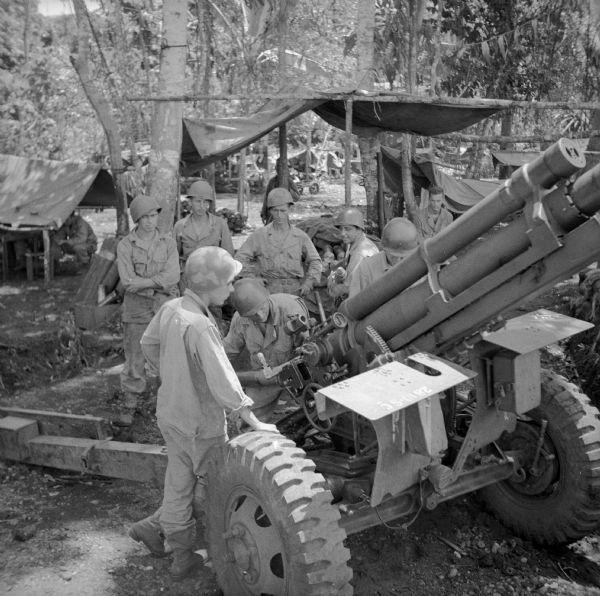 Slightly elevated view of the crew of a 105 mm gun at Saidor, New Guinea (present day Papua New Guinea). The soldier with his back to the camera, at the range finder, is Private First Class John Putnam of Shelby, North Carolina. The soldier loading a shell into the breach is Private Louis Katz of Brooklyn, New York. The soldier opposite Putnam at the gun site is Corporal Willie Fisher of Salem, Oregon. Holding the next shell is Private Anthony De Marco of of Brooklyn, New York. Standing (left to right) behind the gun is Private First Class Harold Schroeder of Bangor, Maine, Private Gilmer Stetzer of Mindoro, Wisconsin and Sergeant Arthur Hoepner of Chippewa Falls, Wisconsin. Trees, tents and more soldiers are in the background.