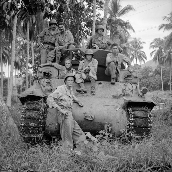 Tank crew at Saidor, New Guinea (present day Papua New Guinea). The soldier's names are, (standing) Sergeant William Preston of San Francisco, California, (hatches, left to right) Corporal Nolan Burns of Milwaukee, Wisconsin, Corporal Earl MacDonald of Alphretta, Georgia, Corporal Harold Kling of St. Paul, Minnesota, (top, left to right) Private Wilber Janes of Des Moines, Iowa, Private First Class Ted Paulos of Vandergrift, Pennsylvania and Private Edward Johnson of Allentown, Pennsylvania. The tank has some art painted on the turret of a snarling black cat inside a star. Foliage is in the foreground and palm trees are in the background.