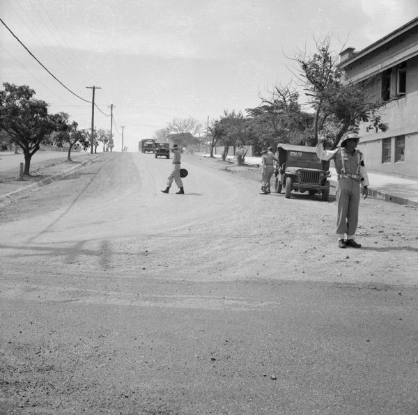 Aussie Military Policeman standing on a Port Moresby main street with his hand raised, New Guinea (present day Papua New Guinea). Jeeps and soldiers are on the street behind him.