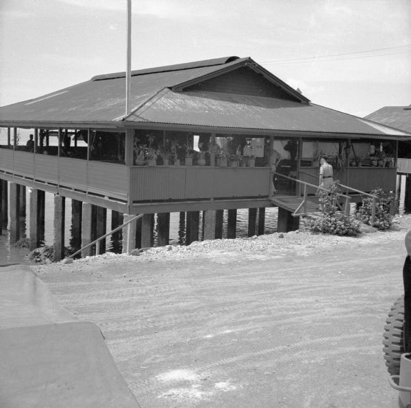 View from road of the exterior of the Allied Officers' Club in Port Moresby, New Guinea (present day Papua New Guinea). It is built on posts above the water on the coast. A large porch or veranda surrounds the interior. Potted plants are lined up on the railing facing the road. An officer is walking up the ramp into the club. Portions of jeeps are in the foreground on the left and right.