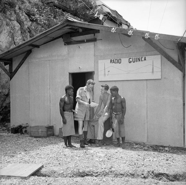 Gordon Williams, of the Australian Broadcasting Corporation, and Stanley J. Quinn of the Mutual Broadcasting System, pose with two indigenous men in front of the Radio Guinea building. For more information about Radio Guinea, see the article in the Clipping Book created by Robert Doyle's mother while he was a war correspondent.