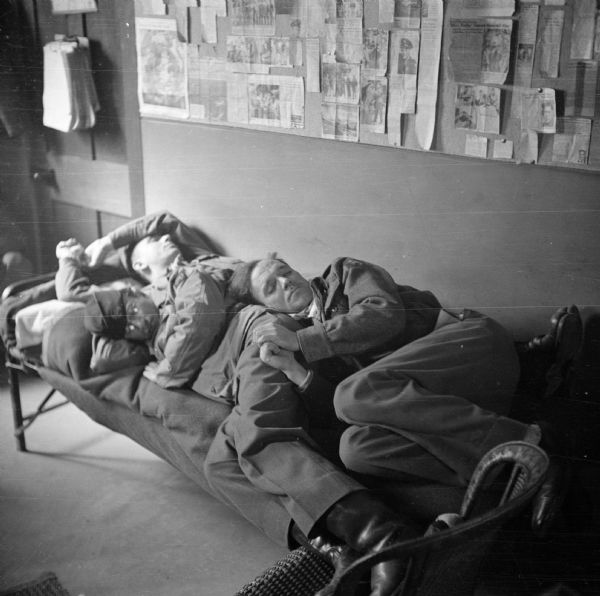 Three war correspondents napping on a cot in the Public Relations office at Earls Colne Airfield, England, on D-Day. Many newspaper articles are pinned on a bulletin board on the wall behind them. Names, (left to right) Collie Small of United Press, Bede Irvin of Associated Press and Bud Hutton of <i>Stars and Stripes</i>, an American newspaper that reported on matters affecting the members of the United States Armed Forces.