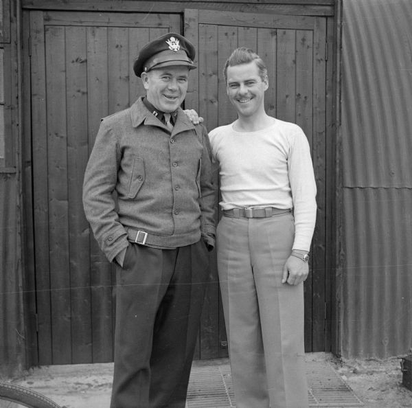 Captain Donald Nicholson of Minneapolis, Minnesota and Major Daniel M. Lewis of Ladysmith, Wisconsin, pose at Royal Air Force station Steeple Morden, located 3.5 miles west of Royston, Hertfordshire, England. They are standing in front of a double wooden door to a metal Quonset hut.