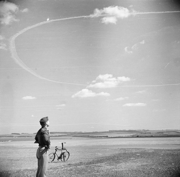 Major Daniel M. Lewis looks up at aircraft contrails at Royal Air Force station Steeple Morden, located 3.5 miles west of Royston, Hertfordshire, England. A bicycle is leaning against a nearby post. Low hills and trees are in the background.