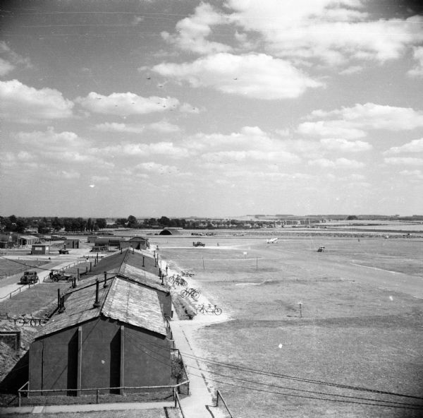 Elevated view of the airfield at Royal Air Force station Steeple Morden, located 3.5 miles west of Royston, Hertfordshire, England. The 355th Fighter Group was stationed there. Bicycles are parked on the right side of the buildings, and planes, trucks and an ambulance are  on the airfield. On the left are several cars. Trees and low hills are in the distance.