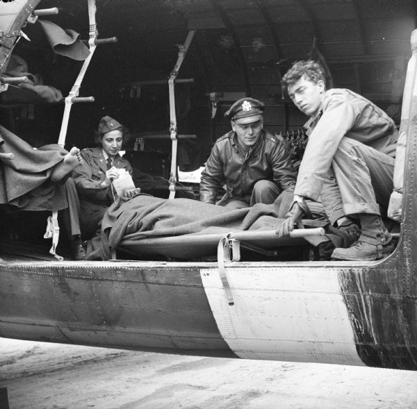 Wisconsin military personnel load a patient covered with a blanket on a stretcher onto a Douglas C-47 Skytrain cargo plane during an air evacuation in Normandy, France. Names, (left to right) are Lieutenant Josephine Sansone Russo of Milwaukee, Captain K.W. Covell, a physician from Racine, and Private James Dietrich of Sheboygan. Robert Doyle, war correspondent for the <i>Milwaukee Journal</i>, made the trip from England with a load of large containers of blood. After the supplies were unloaded, they filled the plane with wounded and flew back to an English hospital. Doyle wrote a caption for this image although it was not published at that time, "Wisconsin nurse and medical soldiers put wounded men aboard an ambulance plane at an airfield on the Normandy beachhead. (See names above.) Russo is stationed in England and flies with planes taking wounded from France. Captain Covell and Private Dietrich are based in Normandy."