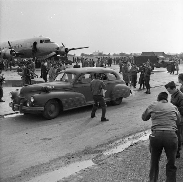 Many photographers take photos of a military automobile leaving an airfield in Paris, France. Robert Doyle notes "arrival of first plane US ATC (United States Air Transport Command) to fly direct from America today." On that plane were General George C. Marshall and Justice James F. Byrnes who were met by General Dwight D. Eisenhower and General George C. Marshall. The plane, on the left, is a Douglas C-54 Skymaster. In the background are jeeps, more airplanes and a group of spectators.