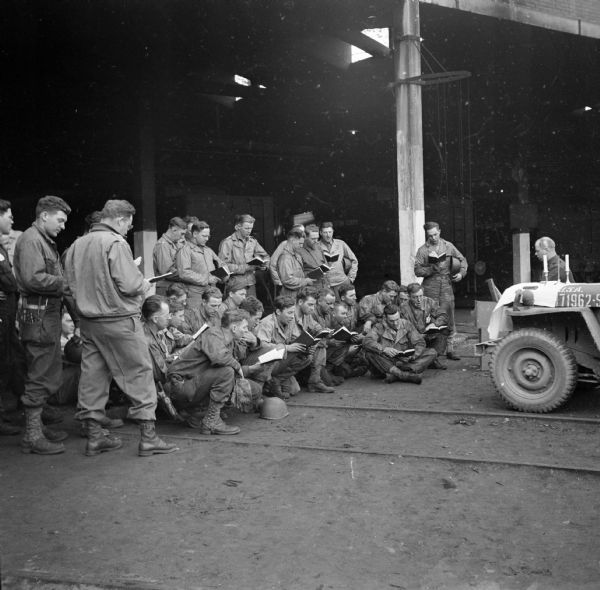 Chaplain Captain Allan R. Fredine holds a Protestant church service in a roundhouse used by the 757th Railway Shop Battalion in Cherbourg, France. He is using the hood of his jeep as an altar and is seated at a portable piano. Many soldiers stand and sit while singing from hymnbooks. Back in the States, he had congregations in Eau Claire and Superior, but more recently his family was in Minneapolis.