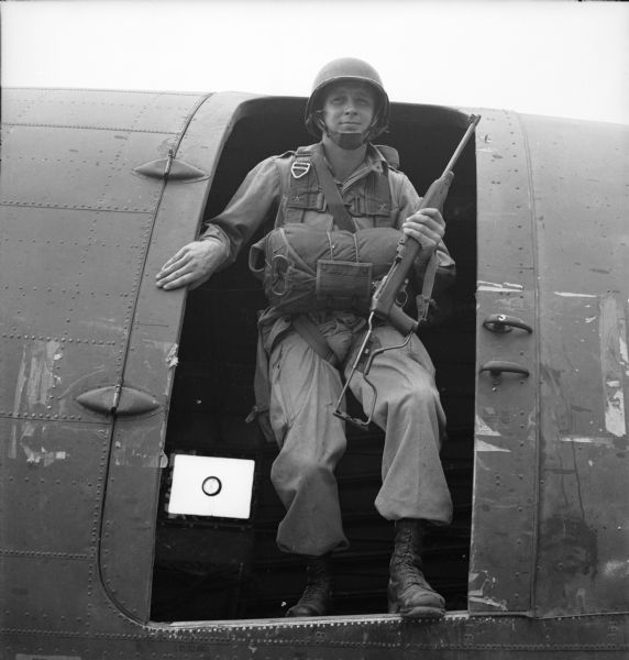 View of a paratrooper just about to jump from the side door of a warplane. He is wearing the full kit and is carrying a rifle. It is likely that this is a posed image for <i>YANK</i>, the Army Weekly magazine. On two sides of the negative is the text, "HANLEY-YANK" and "Pvt. Johnny Jump."