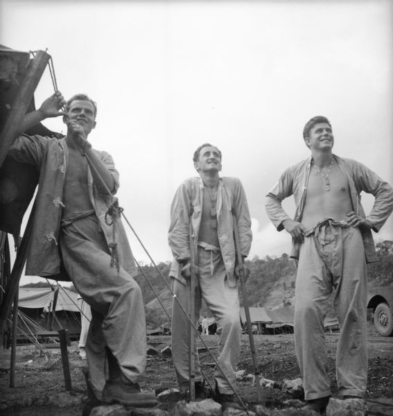 Three soldiers in hospital pajamas gaze up at the sky. The middle soldier is on crutches, and the soldier on the left leans on a tent rope. A jeep, more tents and tree-covered hills are in the background.