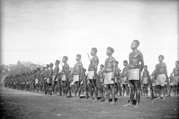 The Royal Papuan Constabulary stands at attention with their weapons in the Port Moresby area, New Guinea (present day Papua New Guinea). The constabulary was a national police force with jurisdiction throughout all of New Guinea. They played a significant role resisting the Japanese occupation during World War II. Their uniforms consist of lava lavas (kilts) and ammunition belts.