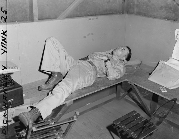 Dick Hanley, of <i>YANK</i> Army Weekly magazine, snapped this photo of Robert Doyle taking a nap on the L-shaped desk in what appears to be a room used by correspondents. Two folding chairs are in the foreground. There is a typewriter on the left with an envelope from <i>LIFE</i> magazine on top of it, and envelopes from the War Department on the right. On the side of the negative is written, "Dick Hanley YANK 25."