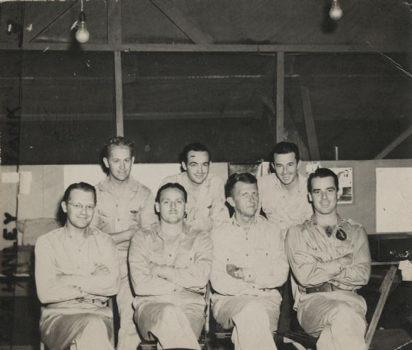 War correspondents pose for a group portrait in the Aussie Press Hut in Port Moresby, New Guinea (present day Papua New Guinea). Names, (front, left to right) Bob Eunson of Associated Press, Dean Schedler of Associated Press, Major T.P. Wagner, Press Officer and Robert Doyle of the <i>Milwaukee Journal</i>, (back, left to right) Sergeant Dick Hanley of <i>YANK</i> Army Weekly, Ralph Boyce of <i>YANK</i> Army Weekly and Lieutenant Joe Jackson, Press Officer. The hut appears to be part tent, part structure. A countertop with a back has equipment stacked on it. Two bare light bulbs hang from the ceiling.