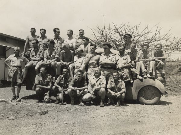 War correspondents, whose typewriters and cameras keep the world informed of action in New Guinea (present day Papua New Guinea), live together in a large hut near Port Moresby. They are posed on or around a military truck. A building and trees are in the background. The complete roster includes (back, left to right) Len Edwards, American Broadcasting Company Technician; Tom Fairhall, <i>Sydney Daily Telegraph</i>; Robert Doyle, <i>Milwaukee Journal</i>; George Moorad, American Red Cross; Reg Leonard, Melbourne Herald; George Silk, Australian Department of Information Photographer; Captain Charles Madden, Australian Imperial Force Conducting Officer; (center, left to right) Harold Guard, United Press; Merlin Spencer, Associated Press; Charles Buttrose, <i>Sydney Morning Herald</i>; Pat Robinson, International News Service, New York; Lewis Sebring, Jr., <i>New York Herald Tribune</i>; Geoffrey Reading, <i>Sydney Daily Mirror</i>; Lieutenant Colonel George Fenton, Australian Imperial Forces Press Relations Officer; Edward C. Widdis, Associated Press Photographer; Martin Barnett, Paramount Newsreel; E.R. Noderer, <i>Chicago Tribune</i>; Dean Schedler, Associated Press; (front, left to right) Chester Wilmot, American Broadcasting Company photographer and British Broadcasting Corporation commentator; Tom Fisher, Australian Imperial Force photographer; George Johnston, <i>Melbourne Argus</i>; Frank Prist, ACME Newspictures, Inc.; Chris Bottomley, Australian Department of Information.
