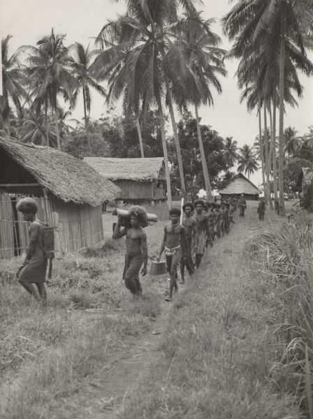 View down line of indigenous men, from a village near the airstrip, helping soldiers carry equipment to a camp area along a trail. Two soldiers stand off to the side in the background on the right. Note how slabs, leaves and vines are used to build the indigenous dwellings.