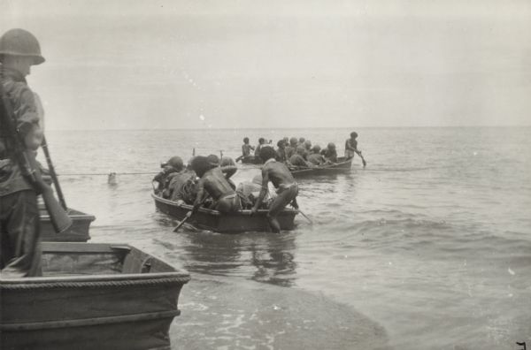Soldiers land on the beach in New Guinea (present day Papua New Guinea). They use collapsible canvas boats to ferry men and equipment from small coastal vessels. Indigenous men help paddle the boats. The accordion-like collapsible panels can be seen on the sterns of two  canvas boats on the left. A soldier with a rifle stands in the boat in the left foreground.