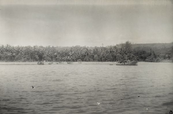 View over water towards the New Guinea coast from a boat. A larger boat is on the right and several smaller craft in the center. A pier is on the shoreline, and soldiers and indigenous people stand on the beach. Supplies are probably being transferred from the larger ship to smaller boats for the trip to a military camp. The jungle and a hill are in the background.
