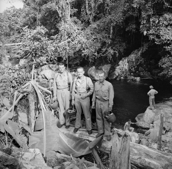 Three officers of General Walter Krueger's staff stand on a log in front of a swimming hole located on Goodenough Island, in the Solomon Sea, New Guinea (present day Papua New Guinea). Robert Doyle describes it, "the natural pool, formed by a cascading mountain stream, is 12 feet deep." A soldier in uniform stands on a rock above the waterline while another nude soldier climbs out below. Doyle also had this to say, "Every afternoon it is filled with shouting soldiers in their birthday suits, seeking relief from the sweltering heat, swimming in the cold, crystal clear water or lounging on the huge rocks." The names of the staff members are listed as Lieutenant Colonel Ray Hamilton (Ham) Martin, Pilot and Senior Aide, of San Antonio, Texas; Lieutenant Calvin Meitz, Co-Pilot, of Burlingame, California; and Captain H.B. (Ben) Dechert, Aide (formerly of the Dallas News), of Dallas, Texas. In his diary, Doyle says that he and the officers went for a swim in the pool. Whether this group portrait was taken before or after their swim is unknown.