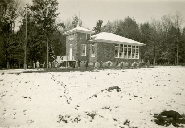 Students at the Cloverland School, District No.2, stand hand in hand, with arms outstretched, in front of the brick school building. The front porch extends upward to form a short tower. There is snow on the ground.